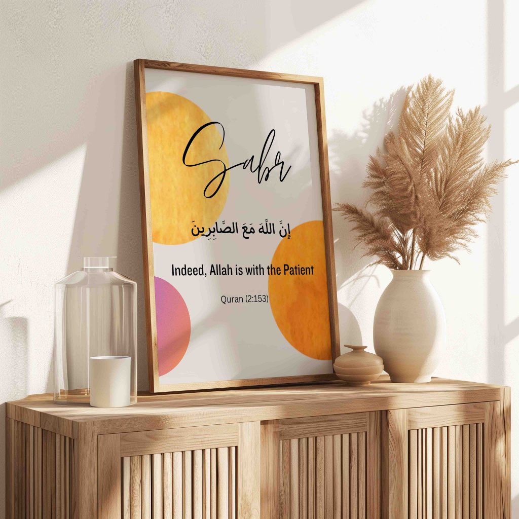 A reminder to practise Sabr: Sabr Arabic calligraphy print with warm yellow, orange and pink circles in a light wooden frame, leaning on a wooden sideboard beside a vase of pampas grass