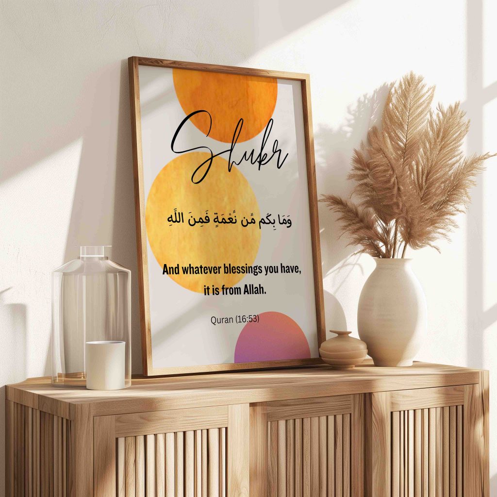 A reminder for daily Shukr: Shukr Arabic calligraphy print with warm yellow, orange and pink circles in a light wooden frame, leaning on a wooden sideboard beside a vase of pampas grass